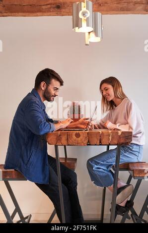 Side view of young Hispanic male freelancer working remotely on laptop sitting at table in kitchen with girlfriend browsing smartphone Stock Photo