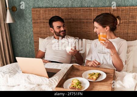 Happy young multiethnic couple in casual clothes having breakfast and watching movie on laptop while sitting in comfortable bed during lazy weekend at Stock Photo