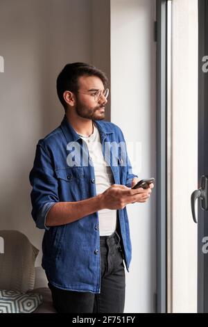 Side view of pensive young bearded ethnic male in casual clothes and eyeglasses standing near window and messaging on mobile phone at home Stock Photo