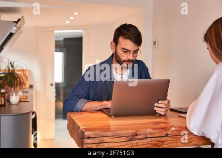 Focused young Hispanic male freelancer working remotely on laptop sitting at table in kitchen with girlfriend browsing smartphone Stock Photo