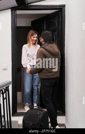 Cheerful young lady meeting anonymous boyfriend with luggage returned from business trip on doorstep of house Stock Photo