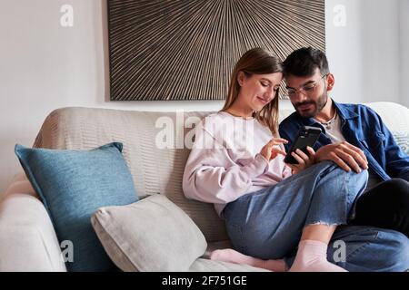 Romantic young diverse couple in casual clothes relaxing on sofa and watching photos on smartphone during weekend at home Stock Photo
