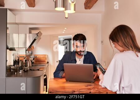 Focused young Hispanic male freelancer working remotely on laptop sitting at table in kitchen with girlfriend browsing smartphone Stock Photo
