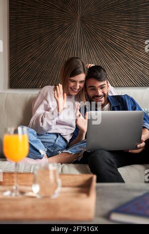 Cheerful young multiracial couple in casual outfits smiling and waving hands while sitting on sofa and having video conversation via laptop Stock Photo