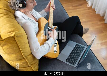 High angle back view of female guitarist in headphones playing musical instrument on couch with netbook at home Stock Photo