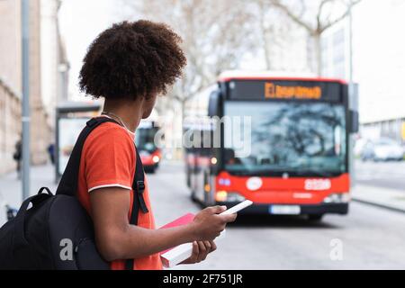 Side view of unrecognizable young African American man in casual clothes and backpack using smartphone while standing on city street and waiting for b Stock Photo