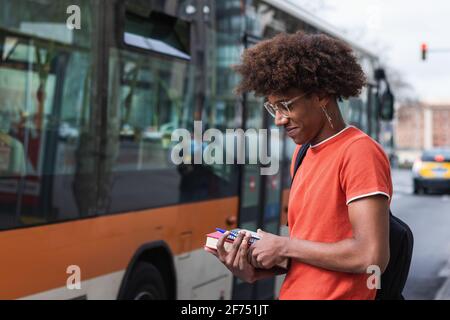 Side view of trendy young black male student with Afro hair in casual clothes and eyeglasses holding books while standing on city street near modern b Stock Photo