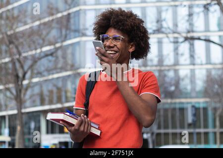 Cheerful young African American male student with curly hair in casual clothes and eyeglasses smiling while using voice message on smartphone on city Stock Photo