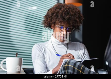 Confident young African American male student with curly hair in stylish outfit and eyeglasses taking notes in copybook while preparing for exam in mo Stock Photo