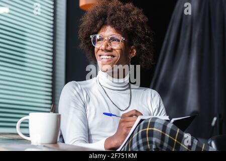 Cheerful young African American male student with curly hair in stylish outfit and eyeglasses taking notes in copybook while preparing for exam in mod Stock Photo