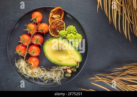 Top view of fresh half of avocado near bunch of cherry tomatoes and sprouts with Romanesco broccoli and nuts on plate Stock Photo