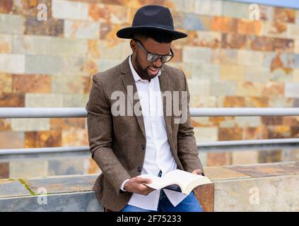 African American male in stylish hat and jacket standing on city street and reading interesting book Stock Photo