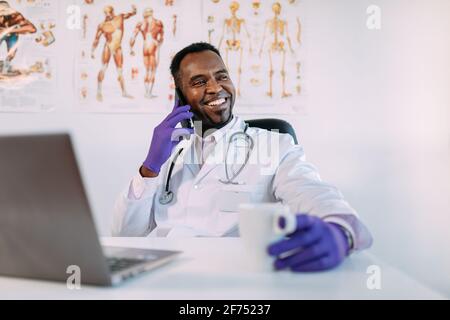 Cheerful young African American male physician in medical apparel and gloves having phone conversation and smiling while sitting at table with laptop Stock Photo