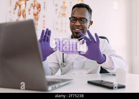 Positive adult African American male doctor in medical uniform and latex gloves smiling and showing mask while having video conference via laptop in m Stock Photo