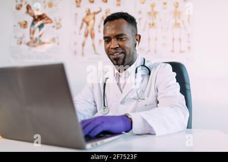 Concentrated young African American male physician in medical robe and TWS earphones working on laptop while sitting at table in modern clinic Stock Photo