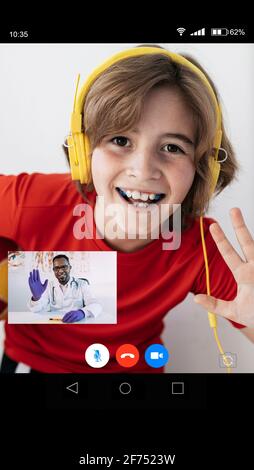 Cheerful boy in casual clothes and headphones waving hand and smiling while talking with African American doctor during online consultation Stock Photo