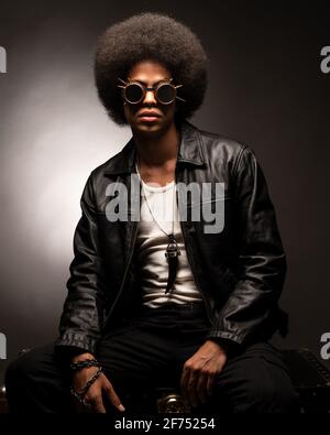 Fashionable African American male in leather jacket and boots sitting on gray background Stock Photo