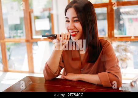 Smiling young Asian woman recording voice note in smartphone while relaxing in modern cafe during break time Stock Photo