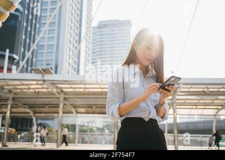Beautiful young Asian woman in formal clothing standing outside office building using smartphone while browsing internet during break Stock Photo