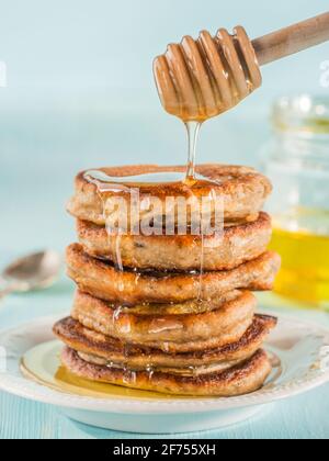 Stack of small pancakes with banana and chia seeds in honey on light blue background. Honey pouring over stack of mini pancakes with chia or poppy seeds. Stock Photo