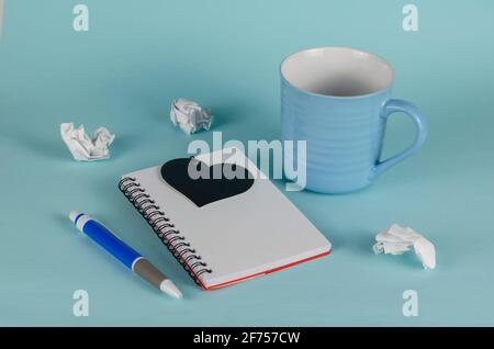 Blue striped mug, pen and open notebook on a blue background. Empty new ceramic mug, black heart symbol and crumpled pieces of white paper. Close-up. Stock Photo