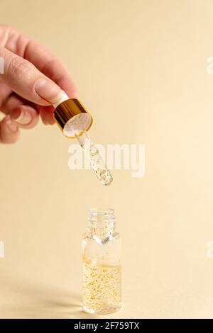 female hand with transparent bottle with beauty serum, hyaluronic acid, 24k gold and vitamins on a beige background. Luxuary home face and body care t Stock Photo