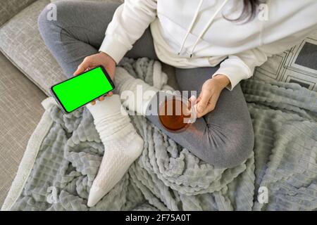 Young person spending time at home using smartphone to chat in sovial network. View from top of bored young woman holding cell with green screen displ Stock Photo