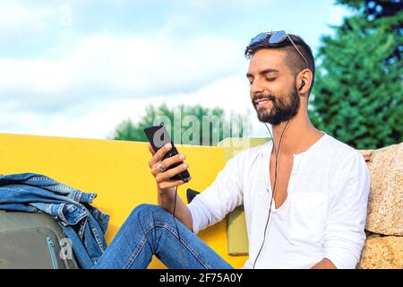 Young solo traveller man sitting on a bench having fun with smartphone listen music in headphones smiling on his own with backpack. New technology tha Stock Photo