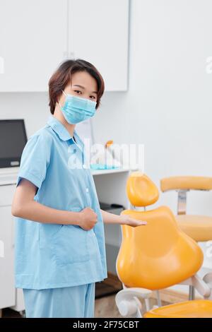 Young nurse in medical mask making welcoming gesture and inviting patient in denal chair Stock Photo