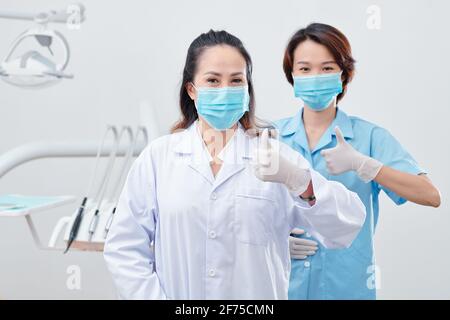 Portrait of cheerful experienced dentist and nurse in protective masks and silicone gloves showing thumbs-up and looking at camera Stock Photo