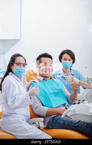 Happy young Asian patient, his dentist and assistant showing thumbs-up and smiling at camera after dental treatment Stock Photo