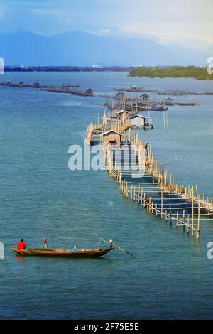 Landscape of fish farming cage in a coastal at sunrise, an Asian senior farmer feeding saltwater fish in fish farming cage. Chanthaburi, Thailand. Stock Photo