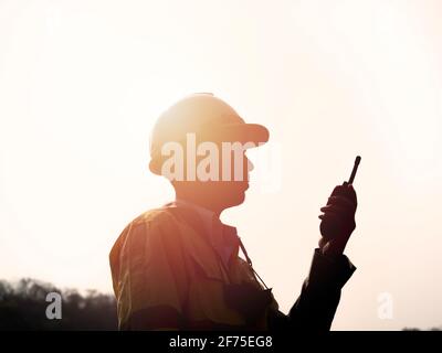 Silhouette side view of young Asian engineering man wearing yellow high visibility safety jacket and white safety helmet or hard hat using walkie talk Stock Photo