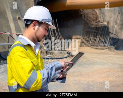 Young handsome Asian tunnel engineering wearing high visibility jacket and white safety helmet working and using digital tablet at tunnel construction Stock Photo