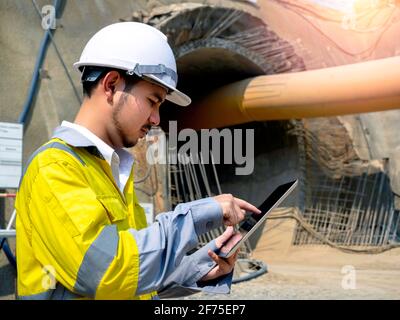 Young handsome Asian tunnel engineering wearing high visibility jacket and white safety helmet working and using digital tablet at tunnel construction Stock Photo