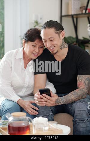 Smiling mixed-race man visiting mother and showing her photos and videos on his smartphone Stock Photo