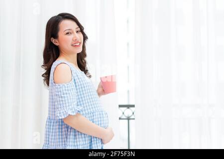 Beautiful woman holding her stomach and holding a cup of milk stands by the window Stock Photo