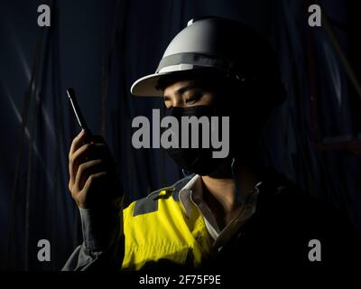 Young Asian engineering man wearing yellow high visibility safety jacket, face mask and white safety helmet or hard hat using walkie talkie in dark ra Stock Photo