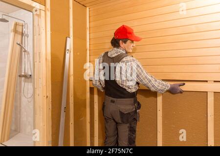 Sauna construction, finishing. The man is screwing a wooden bench to the wall. Stock Photo