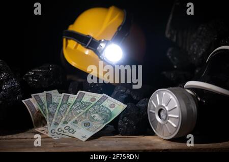 A protective helmet with a glowing flashlight and a dust mask with a filter and paper bills lie on top of the black coal Stock Photo