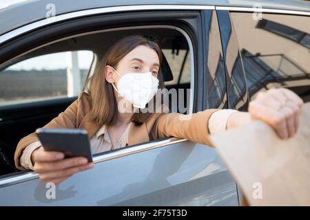 Young woman with protective mask on her face takes the order to go from the car. Contactless payment and self-pickup. Healthcare, virus protection concept.  Stock Photo