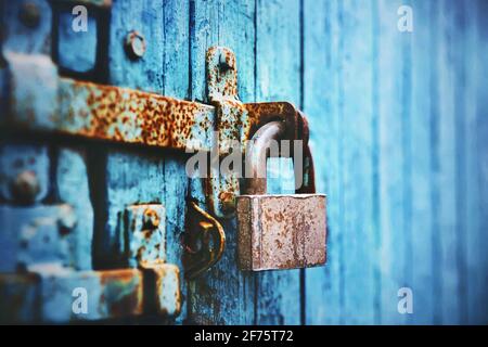 An old large lock hangs on a rust-covered blue abandoned wooden gate. Closed. Stock Photo