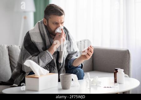 Man covered in blanket sneezing nose and checking for fever Stock Photo