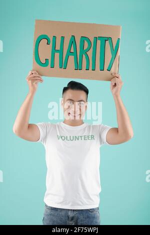 Portrait of handsome smiling young Asian man holding placard with charity inscription and looking at camera Stock Photo