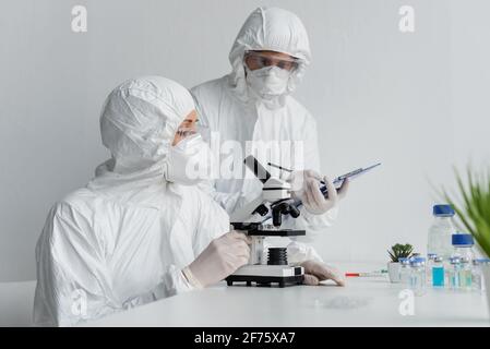 Scientist using microscope near colleague with clipboard and vaccines Stock Photo