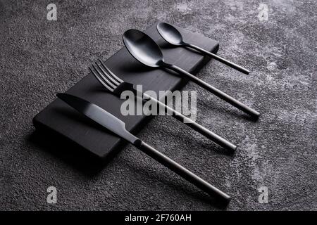set of black knife, fork, spoons on a wooden base. Dark minimalistic still life of exclusive restaurant tableware, concrete background. Stock Photo