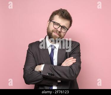 Friendly Caucasian bearded man in glasses and a suit stands with crossed arms and smiles cutely. Pink background. Stock Photo