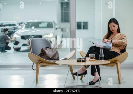 Asian customer woman reading and waiting mechanic checking for maintain her car in maintainance service center with the part of showroom Stock Photo