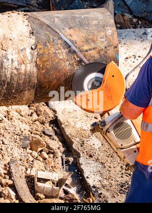 Worker in protective wearing cutting large metal pipe with industrial grinder in  trench Stock Photo
