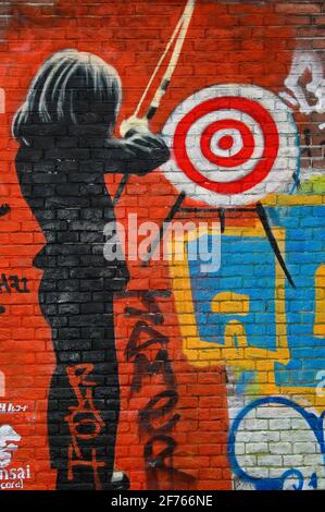 Taking aim … a female archer wearing a dark suit prepares to fire an arrow at a target on a brick wall sprayed bright red in psychedelic street art on one of the former long-term squatters’ strongholds close to the world-famous Slangenhuis or Snake House in Spuistraat, Amsterdam, North Holland, Netherlands. Stock Photo